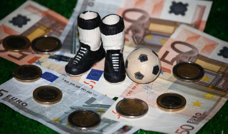 Ein Symbolbild. Die Miniaturmodelle von Schuhen und einem Fußball liegen auf Geldscheinen.