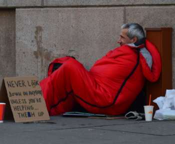 Ein Obdachloser in einem Schlafsack liegt gegen eine Wand gelehnt auf der Straße.